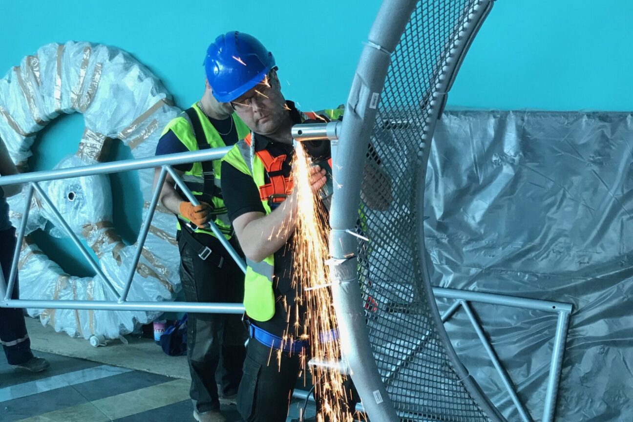 A builder in a green high-visibility jacket, glasses and a blue hard hat is working on one of the metal wheels, bright orange sparks are flying downwards from his tool.