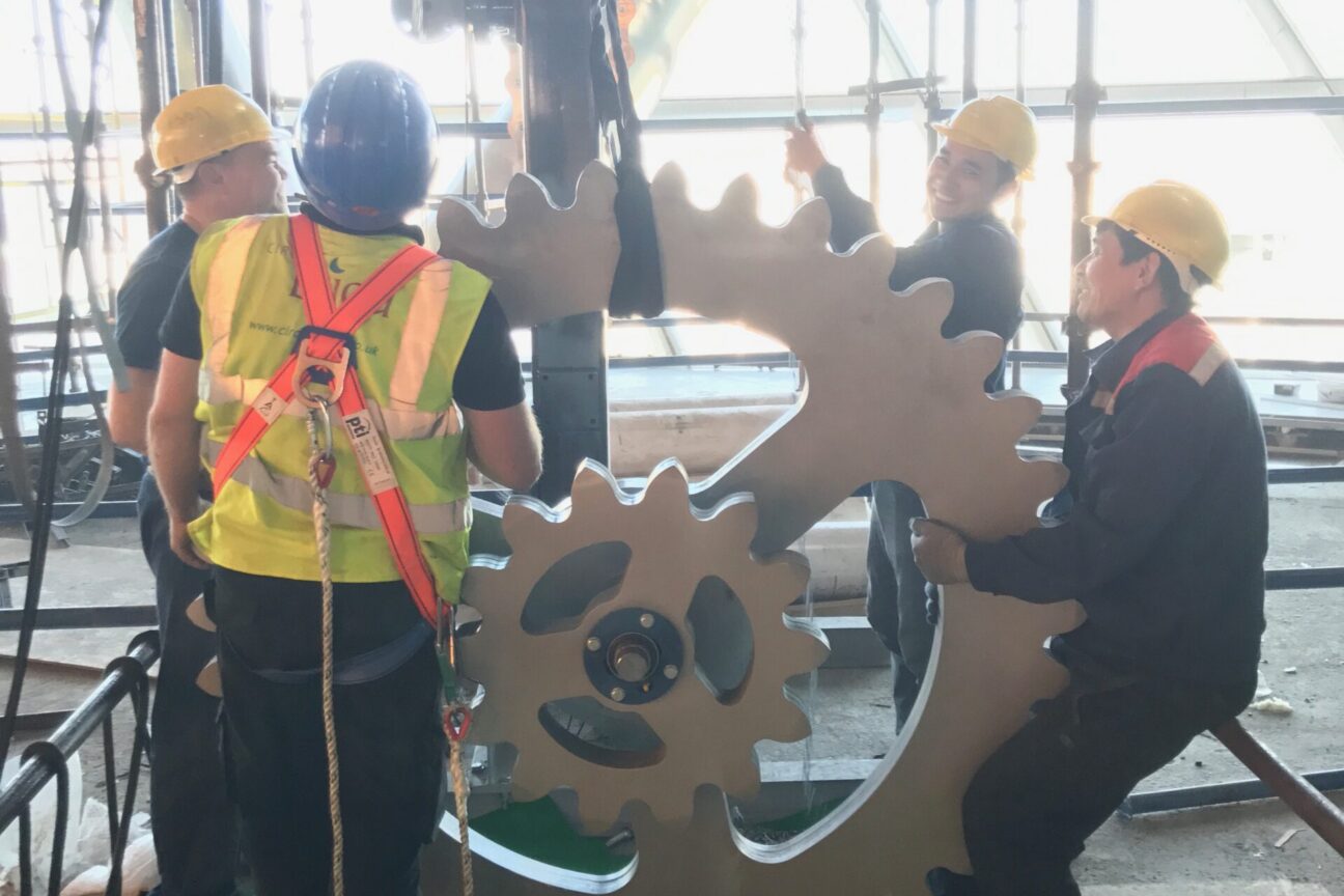 Four crew members with Cirque Bijou high-visibility jackets and hard hats are holding a large grey cog as part of the set for 'Wheel of Energy'. One of the builders is smiling towards the cameras whilst the others are looking up and at the piece of set.