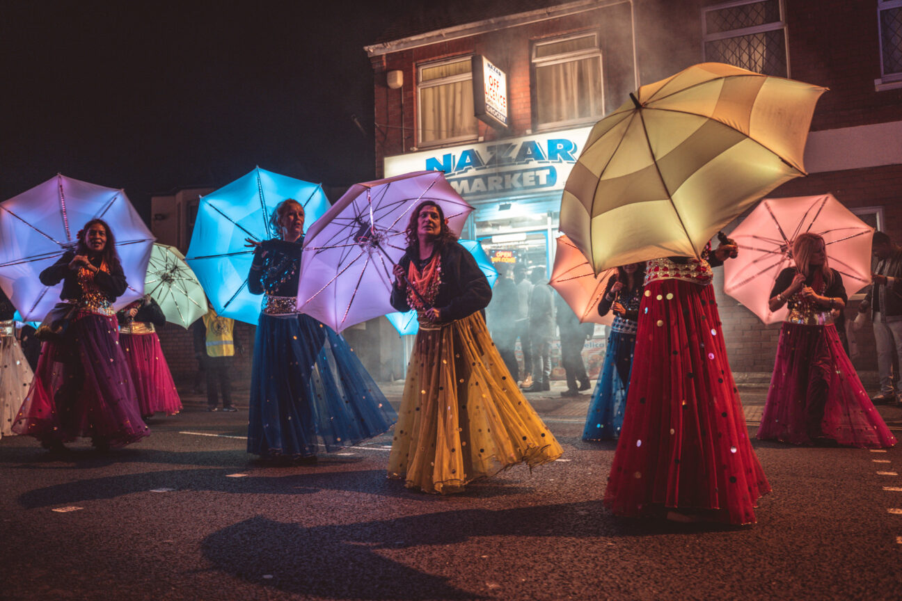 Coventry carnival of lights, female performers in blue, red and yellow skirts hold their LED umbrellas back behind their heads.