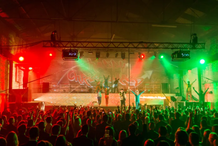 Project 3Sixty. An audience look onto a stage with the writing "Happy Christmas" projected onto a brick wall backdrop. A male performer stands with a mic as a pole performer in all green puts her arms up to bow.