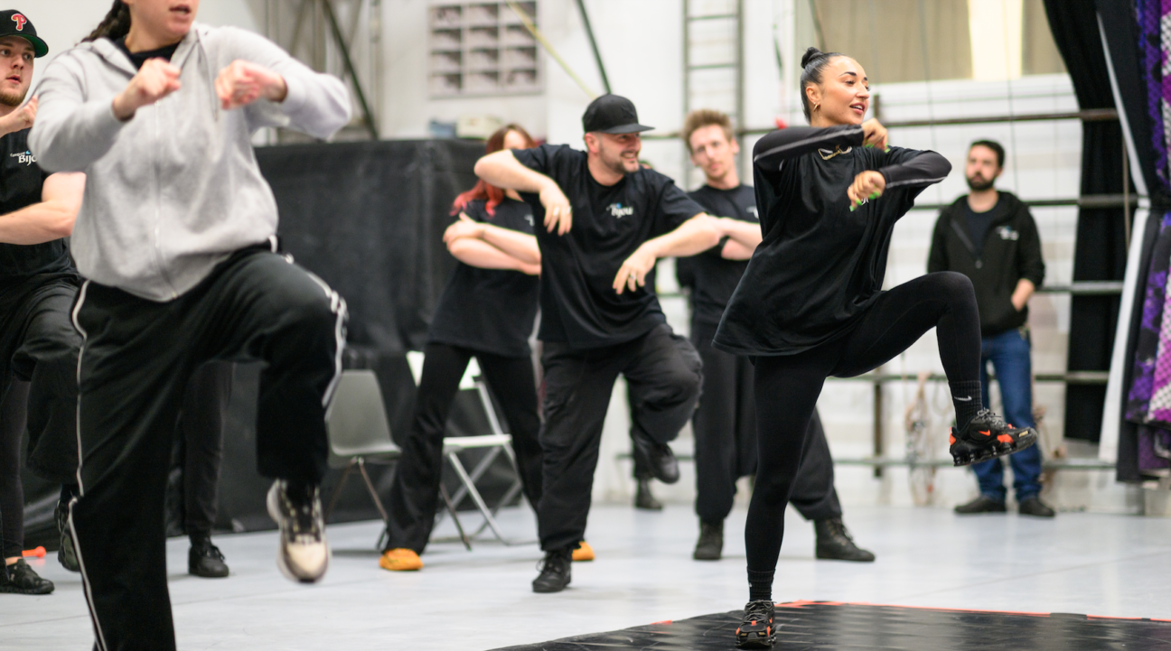 A rehearsal shot of 7 performers practicing a dance routine/ A female leads at the front with her arms and one leg raised, other performers copy behind.