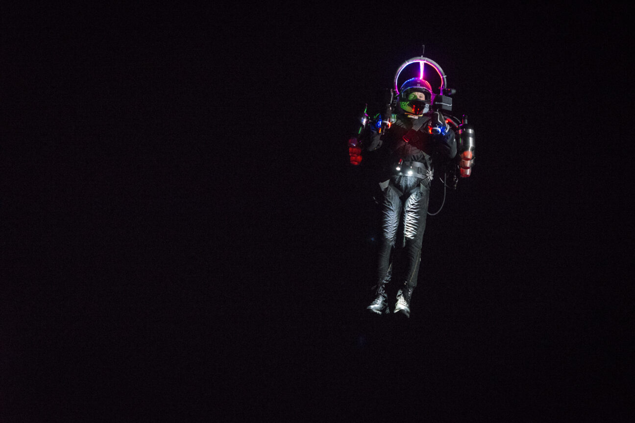 With a black background, a man in a spacesuit with a rocket backpack hovers to the right of the background with a helmet reflecting colourful LED light from a ring above his head. He has two torches around his waistband pointing downwards lighting his black shoes.