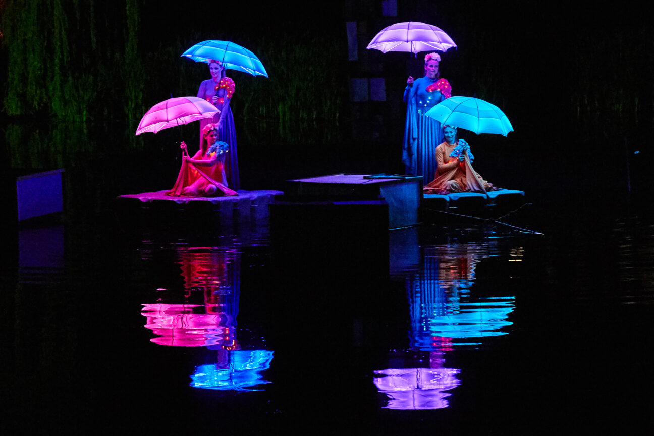 Cirque Bijou, Shambala 2017. Four ladies, (two sitting, two standing), holding pink, blue and purple LED umbrellas that reflect off of the water in front of them.