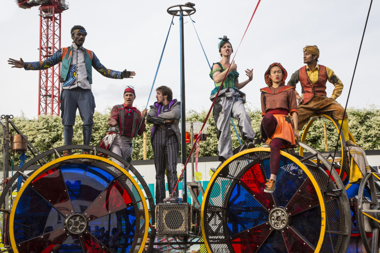 6 performers are stood and sat around 2 colourful wheels. They are costumed in bright coloured, suits, corsets, waistcoats and trousers. A female performer in a green corset looks to be speaking whilst gesturing towards herself, as the other performers look out and at each other.