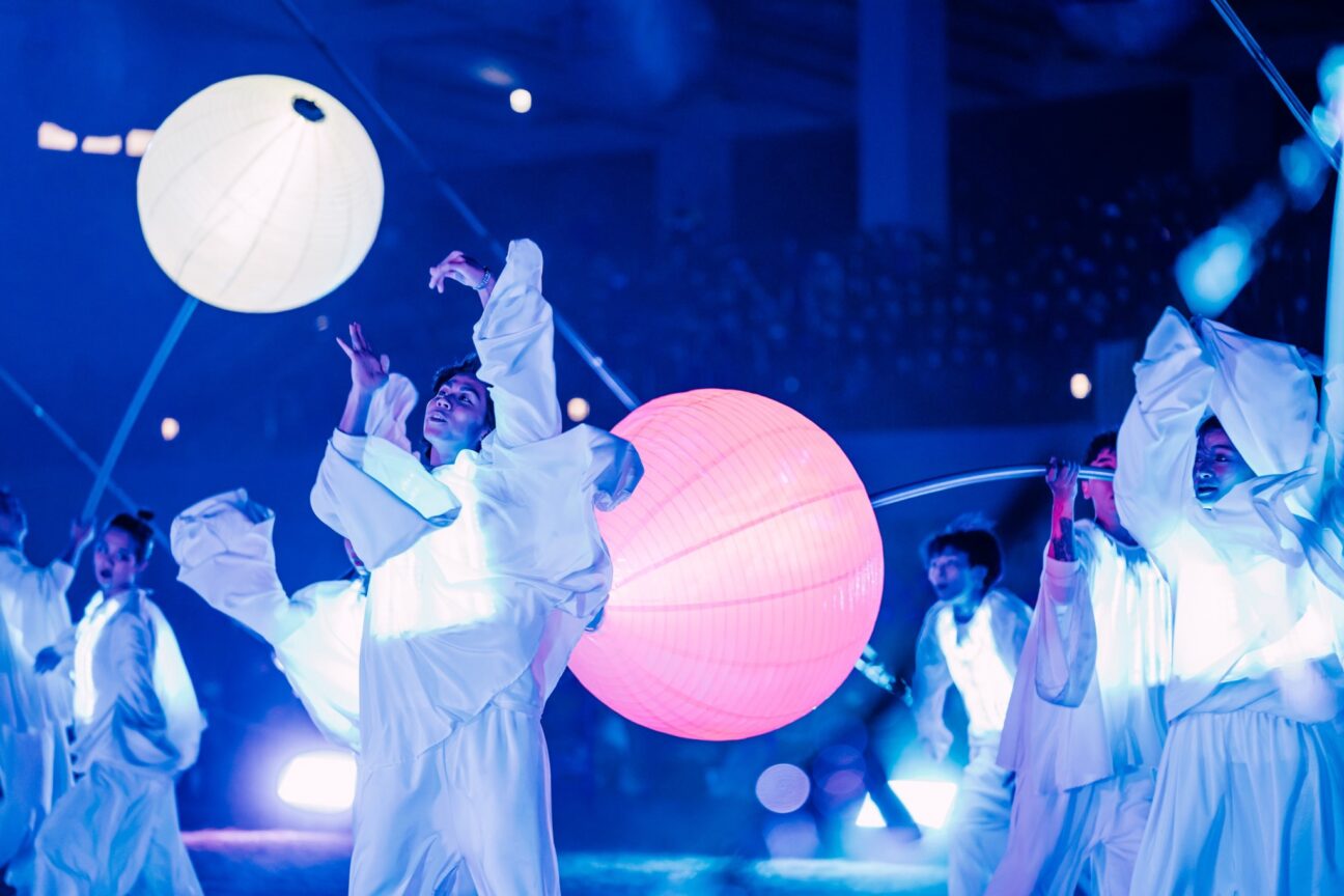 Taiwan Lantern festival, performers are wearing white costumes that are lit with white light. A performer centre of the image is focusing on the movement of his hands above his head whilst other performers are holding and manipulating large pink and white, circular lanterns. Behind the performers is a blurred audience in a dark blue light.