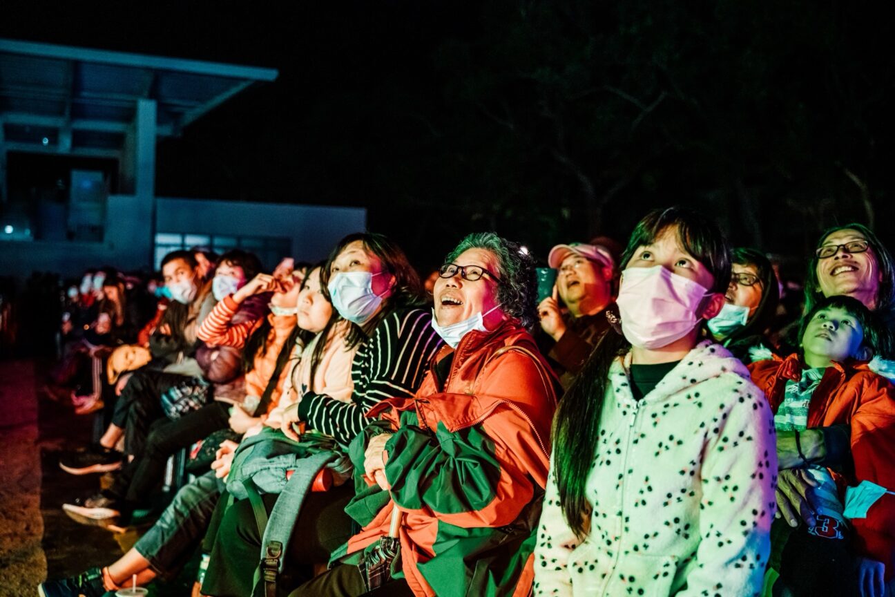 Taiwan Lantern Festival audience are sat gazing upwards, wide-eyed. Some of the audience members are wearing face masks. A lady in the front row, wearing glasses smiles at the lanterns in amazement.