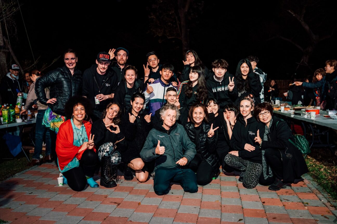 Billy Alwen poses in the centre, on the floor with his thumbs up, in front of performers and crew of the Taiwan Lantern Festival 'Balance of the Cosmos'. The performers and crew are wearing a mix of black clothes, Cirque Bijou tops bar one lady, bottom left, who is wearing a blue shirt and a red jacket. The performers and crew are making peace signs and love heart gestures with big smiles.