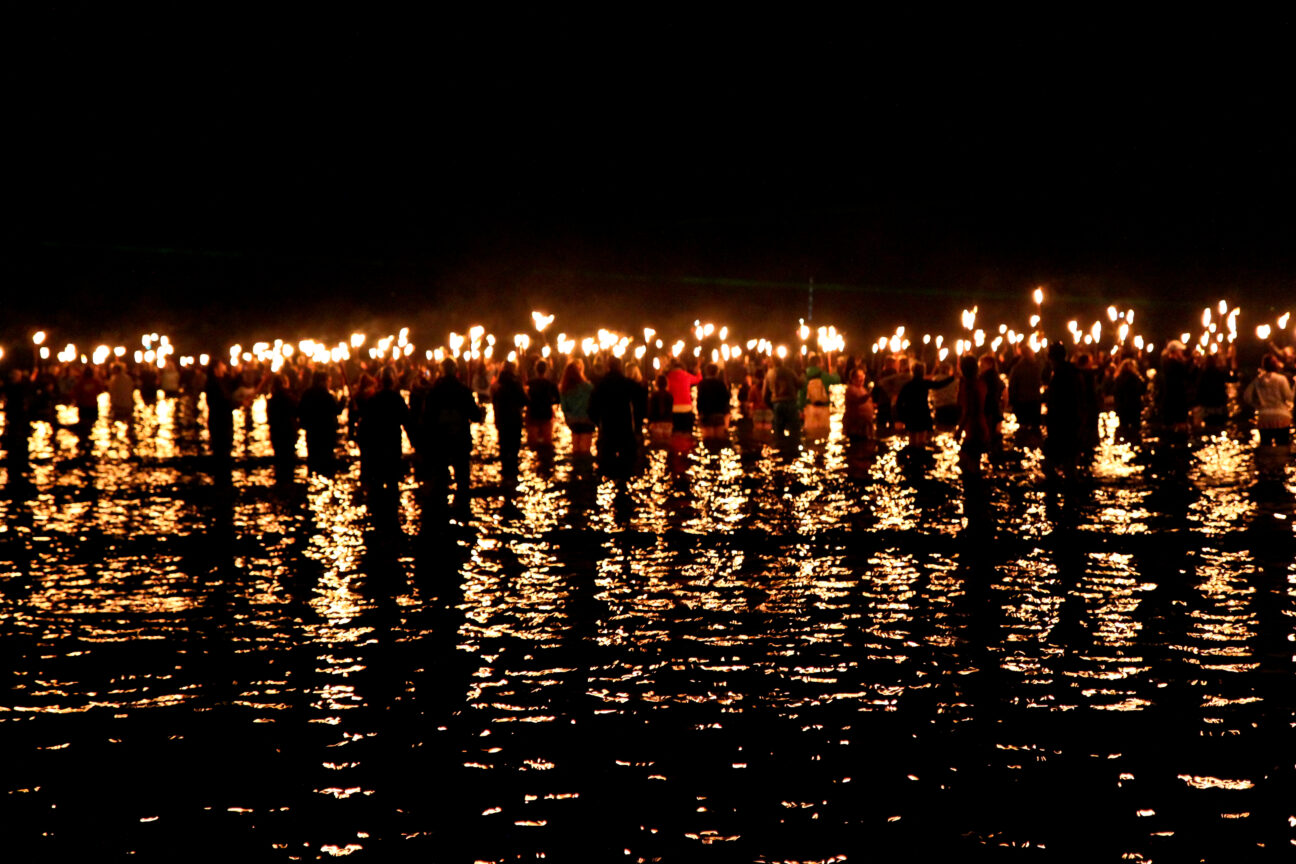 Battle for the Winds, 2012. A group of people in the water marching away from the camera with fire torches raised to the sky.