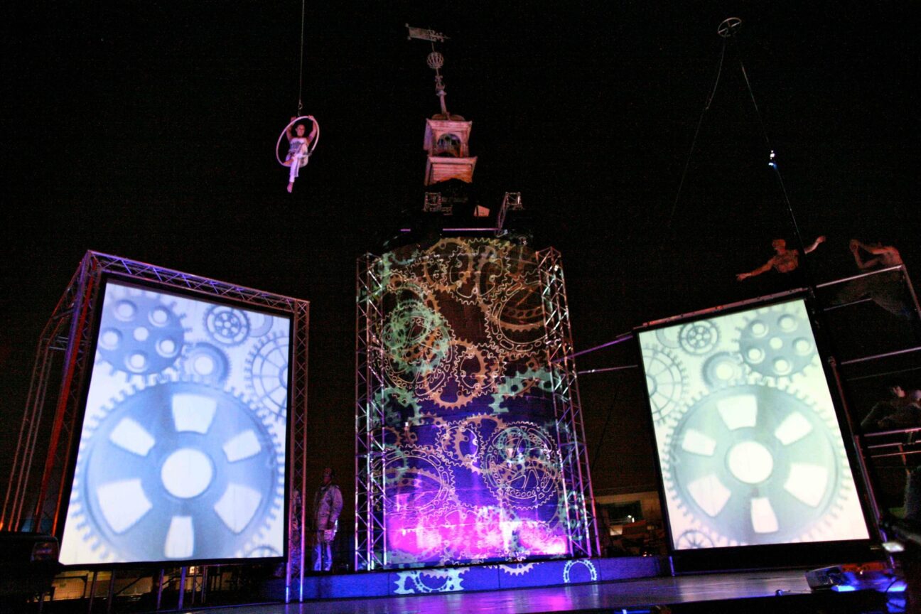 Project 3Sixty. A performer sits top left of the image, cross-legged and with their hands on top of the aerial hoop that is suspended high in the air. There are images of cogs stage left and stage right on screens and a projection of green and orange cogs upstage centre.