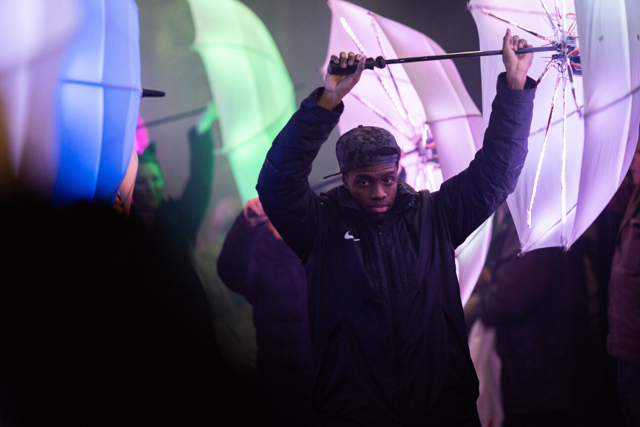 Male performer holds purple LED umbrella out to his right side.