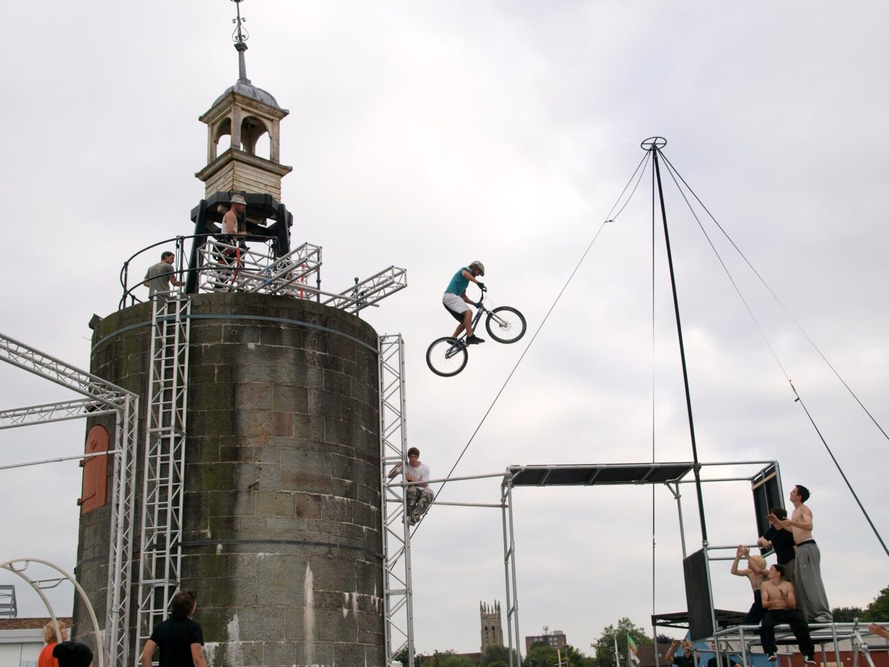 A performer on a bike is captured mid flight between structure and platform. Fellow cast and crew look on at him in supsense.