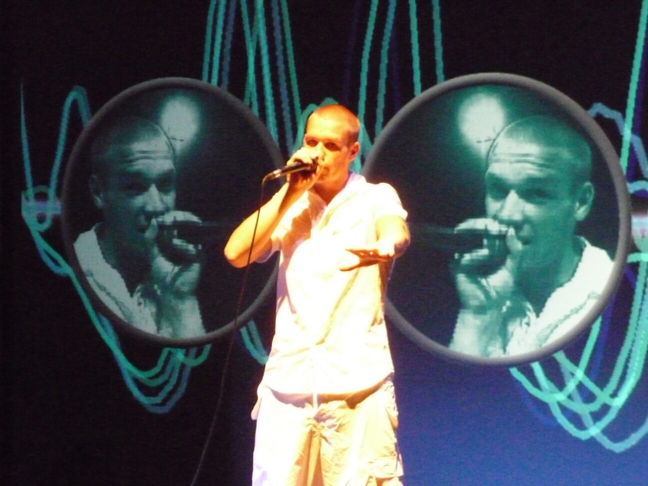 A performer stands centre of the image, dressed in white holding a hand mic to his mouth. behind him, either side he is projected onto circular screens where there are now three images of him.