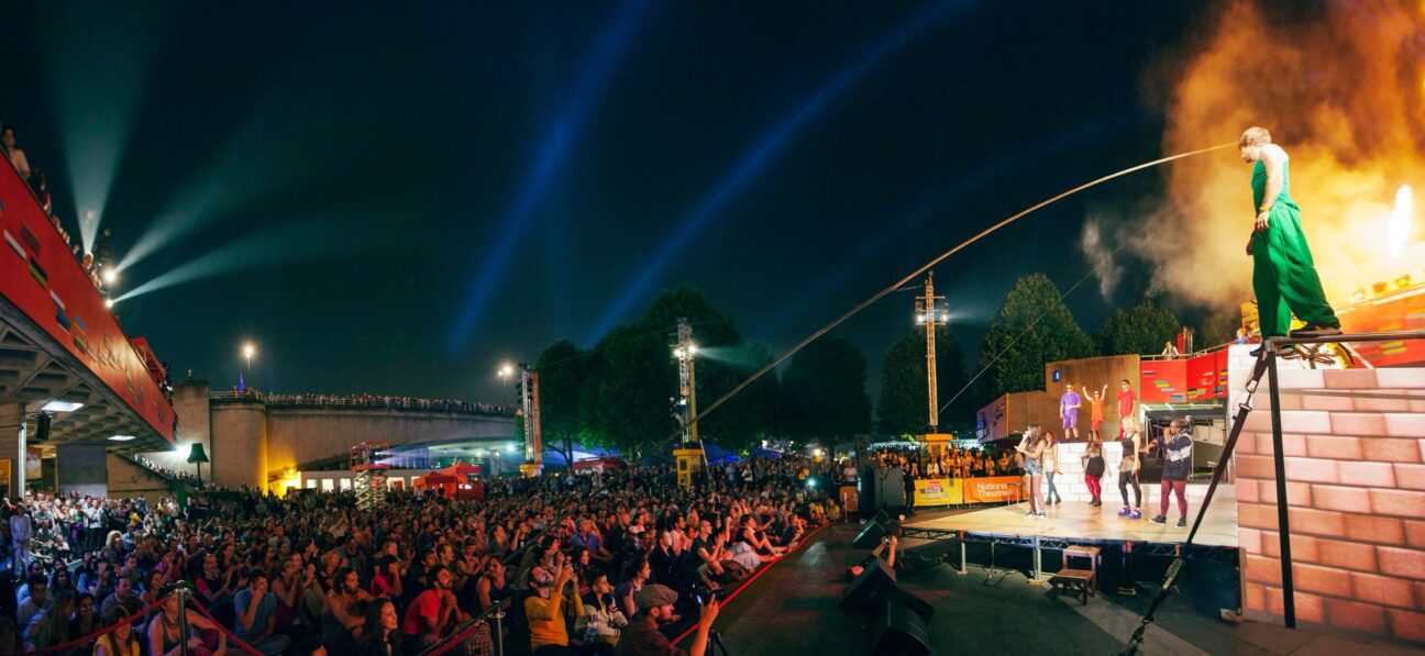 Project 3Sixty. A large audience looks towards the stage where there are 9 performers dressed colourfully. Two have mics in their hands and others are dancing around the, They are stood on a slightly raised stage with a "brick-wall" platform to the right of the image, of which, a performer dressed in all-green, stands on top of.