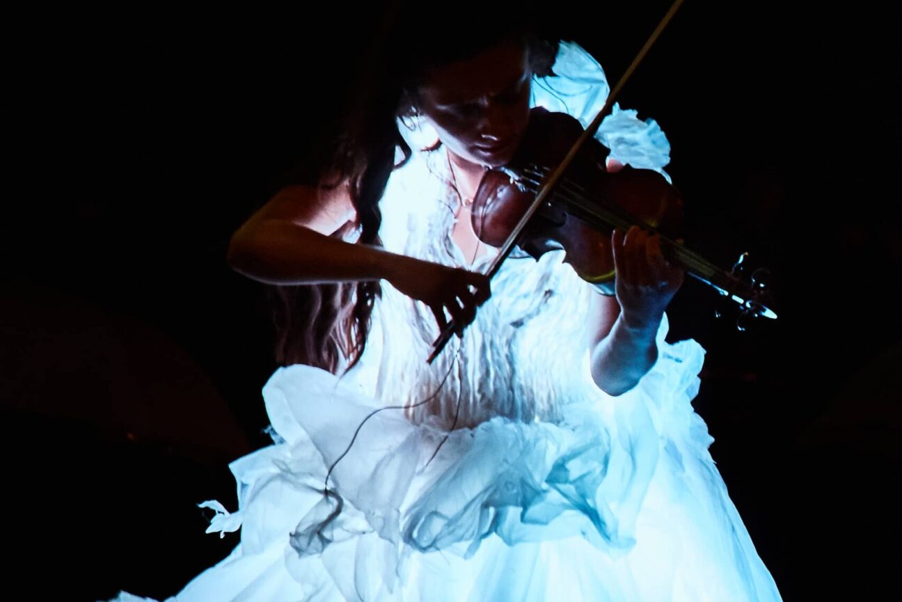 A girl plays the violin in a led lit dress against a dark background