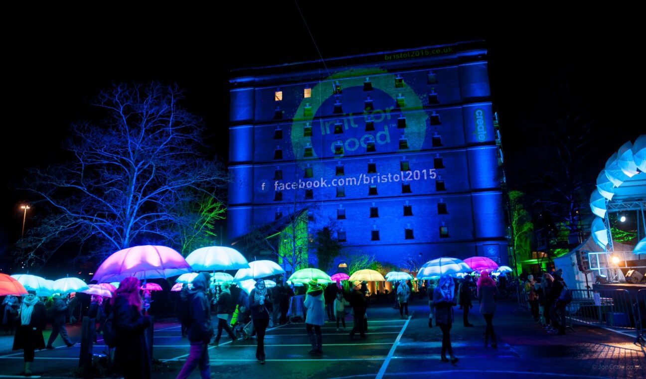 LED umbrella performers infront of an industrial building with projections on