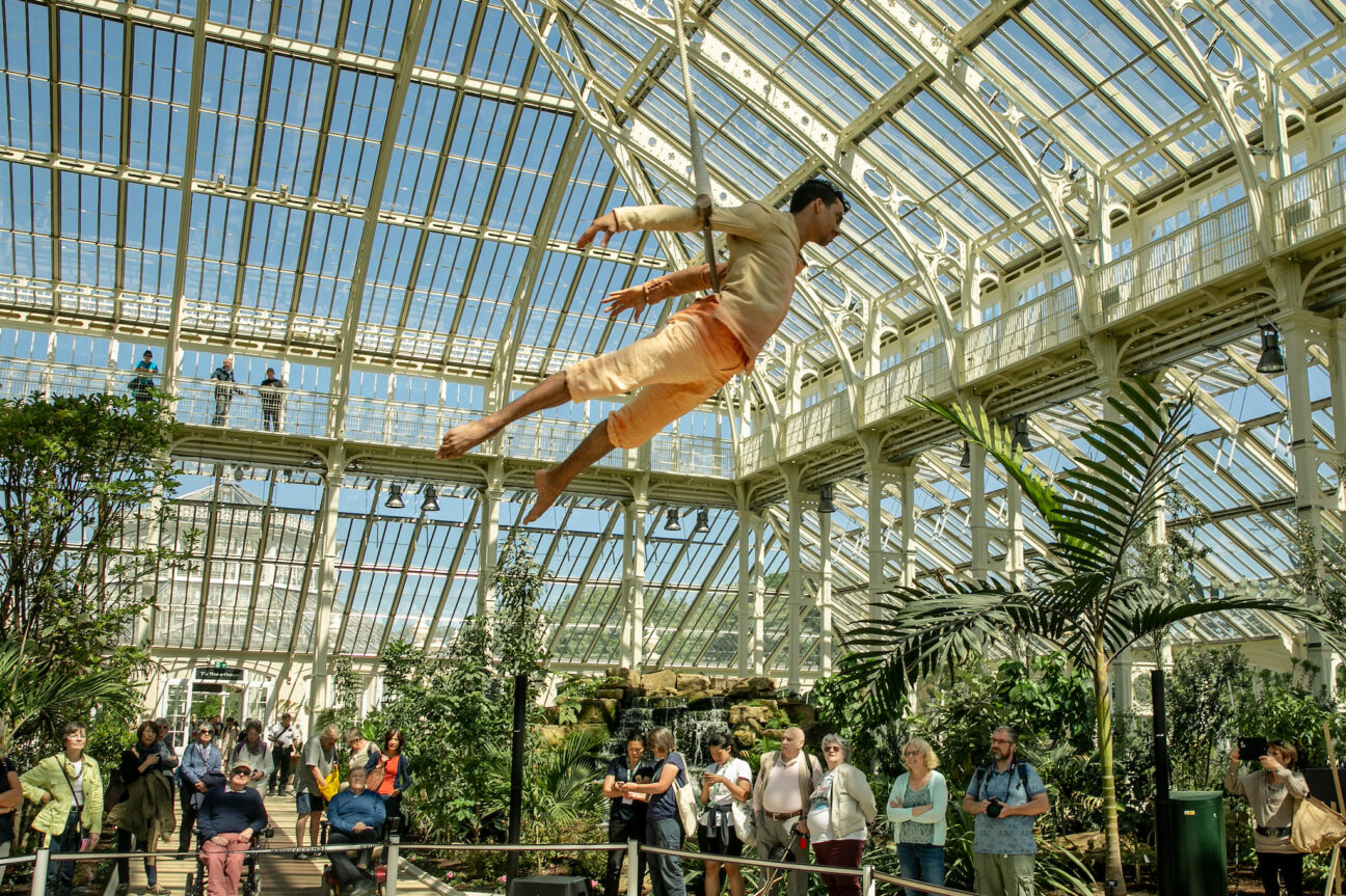 Kew Gardens. Performer, Korri, is suspended above an audience who look up in amazement, some taking photos. He is surrounded by trees and there is a water feature in the background of the image.