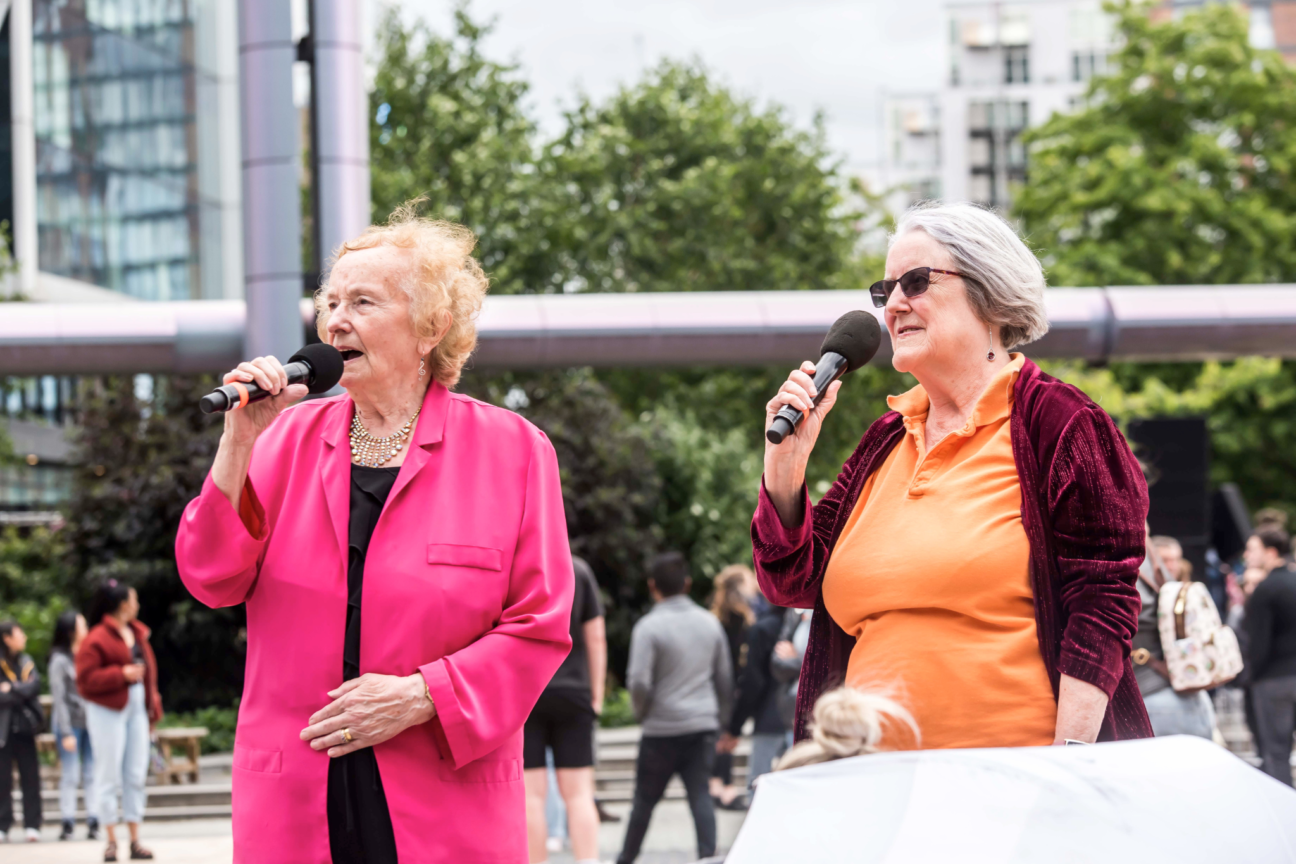 Two female singers dressed in pink and orange, both singing into black microphones.