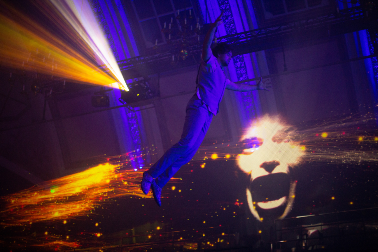 Voodoo Halloween male performer opens his arms out as he hangs in the air with orange lighting behind him and a projection of a screaming face to his right.