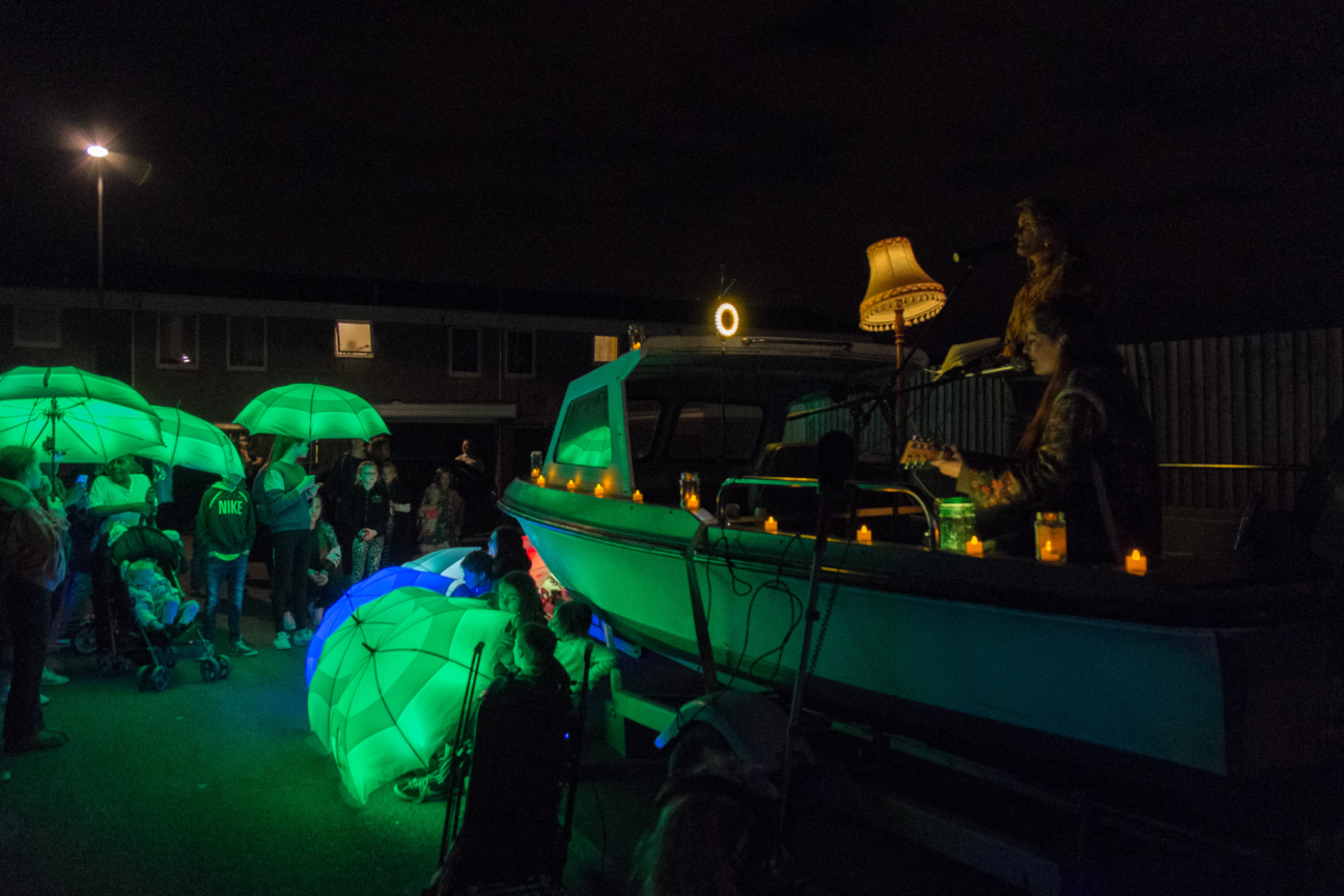 A guitarist and singer sits in a dimly lit boat surrounded by LED umbrellas outside the boat and held by audience members. In the boat there is a lampshade and tealight candles.