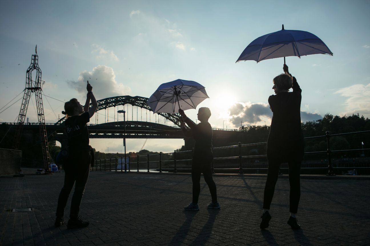 2 women raise umbrellas up outside at Sunderland Tall Ships. There's a Cirque Bijou member directing them.