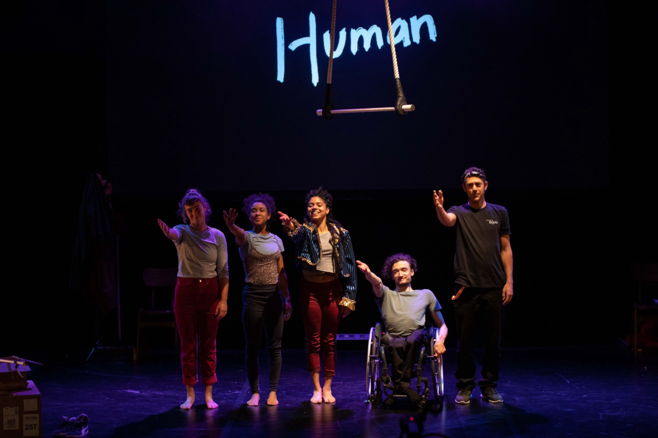 Human, 5 performers gesture out to thank someone after their performance. There is a trapeze hanging above them, and Human projected in blue as a backdrop.