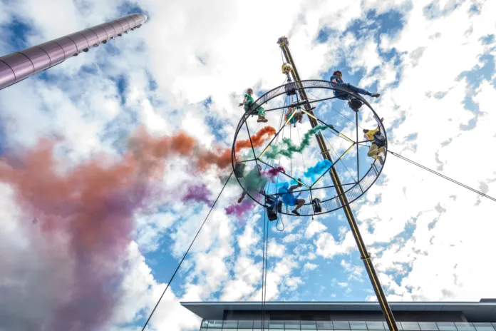 Way Above the Houses. 6 performers sit on top of the aerial carousel with coloured smoke released travelling backwards. There is a blue sky above and one of the performers is looking own towards the camera waving.