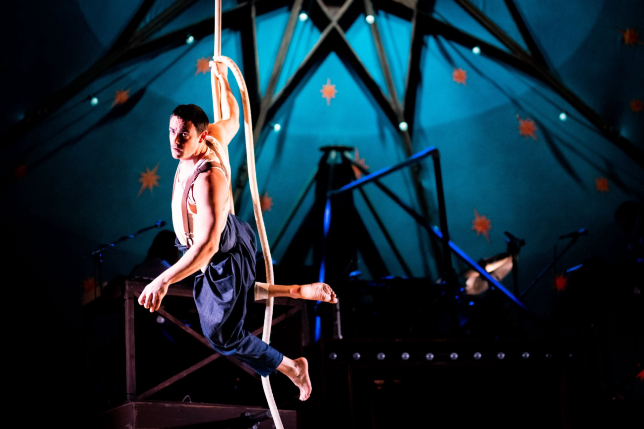 A male performer looks out towards the audience, he has one hand on the rope and the other free as he suspends in the air.