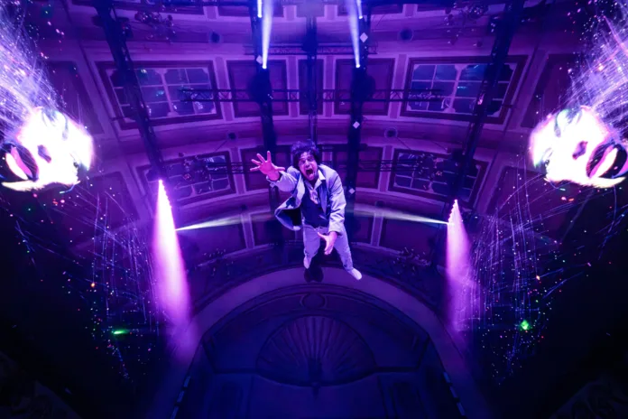 Voodoo Halloween, a performer hangs down facing the camera making a scared expression with his hand reached out towards the camera. He is wearing a suit and there is purple lighting above him.