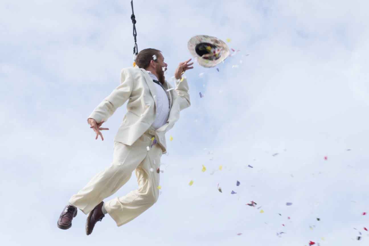 Jamie Beddard in Weighting. Performer Jamie is suspended from a crane in a cream suit.