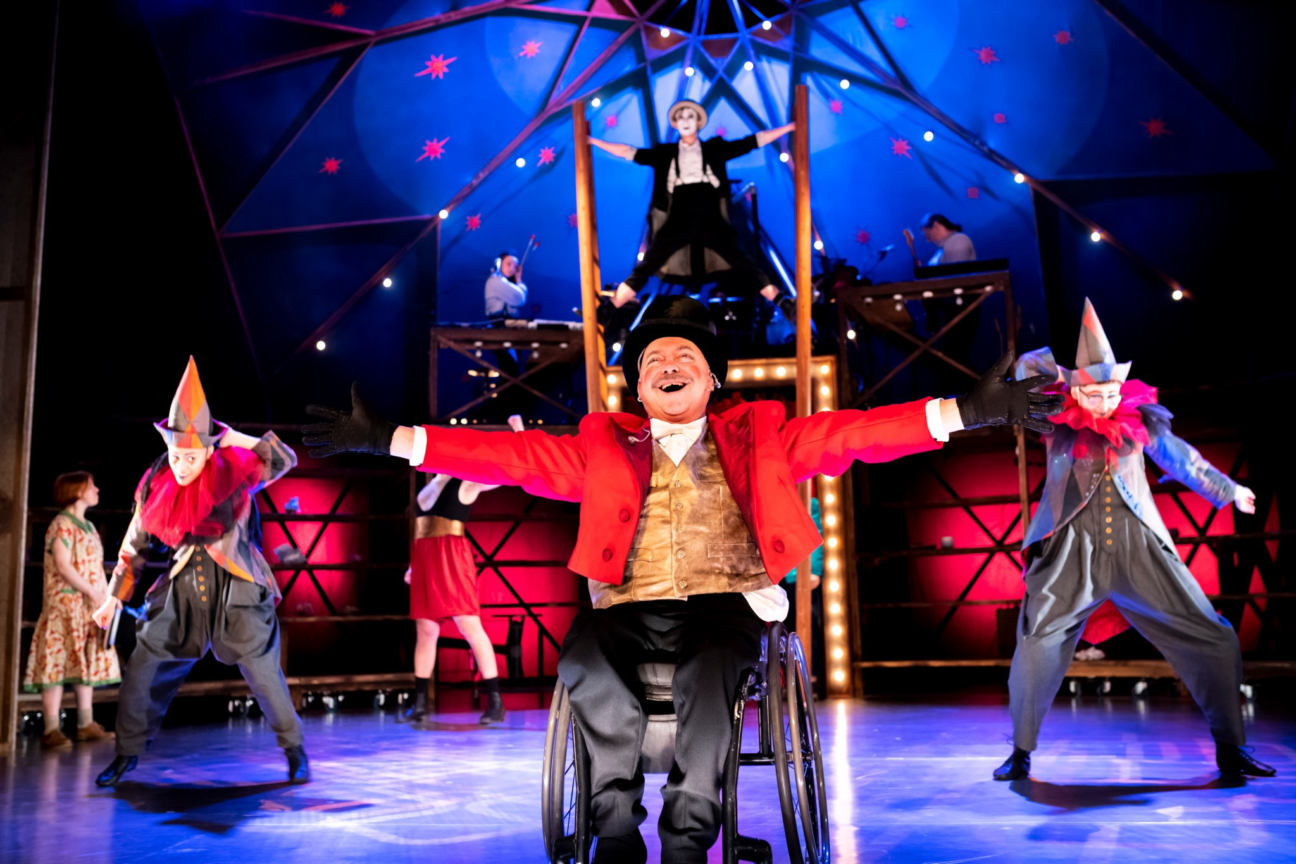 Waldo's Circus of Magic & Terror. A ringmaster performer, wheelchair user, is centre of the image with his arms out wide and a large smile. Behind him are two clowns in grey and red costumes with their arms raised. You can see the big top in the background and there is a performer stood between two ladders above the ringmaster.