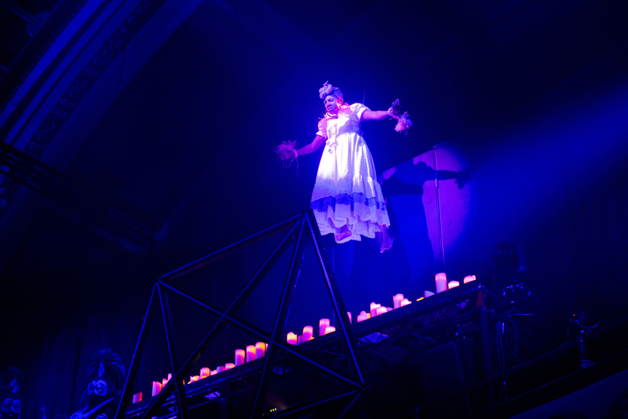 Voodoo Halloween female performer in white frilly dress and purple turban is suspended in the air over lit candles, looking out over the audience.