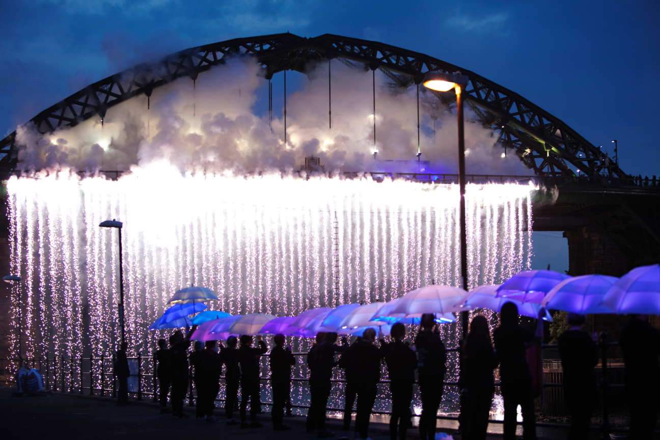 People stand with LED umbrellas to the side of a pyrotechnics waterfall from the bridge.