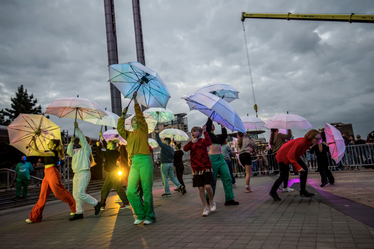 Way Above the Houses. Young people in bright coloured clothing hold up coloured umbrellas facing outwards in different directions. There is a bright orange light shining through the bottom of the image.