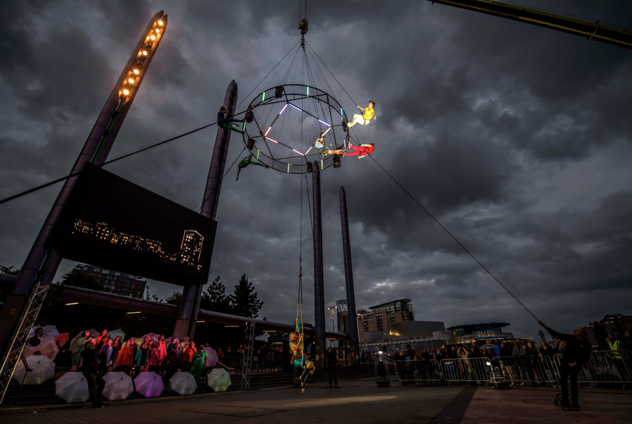 Way Above the Houses. 4 performers move around an aerial carousel suspended in the air. One performer hangs on an aerial hoop lower to the ground. Behind the performer is a choir dressed in colourful clothing with umbrellas around them. The aerial carousel has neon lights on it.