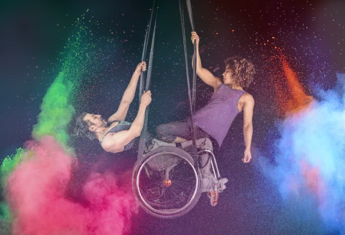 Two performers are suspended in the air holding onto two black straps. One performer is using a wheelchair and the other performer is facing him sat on his knees. There is an explosion of paint either side of the performers.