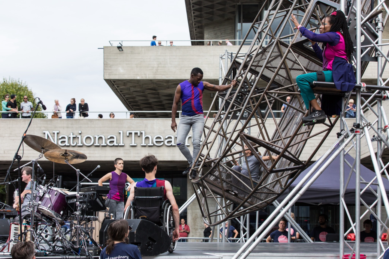 Outside the National Theatre, performers are looking into metal wheel structure where a female performer, in grey, is sat inside. There is a band in front of the wheel with a drum kit and piano.