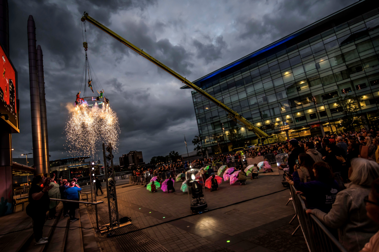 Pyrotechnics releases from the aerial carousel at Way Above the Houses. Performers crouched with umbrellas over their heads below the carousel. The audience watch on form a distance.