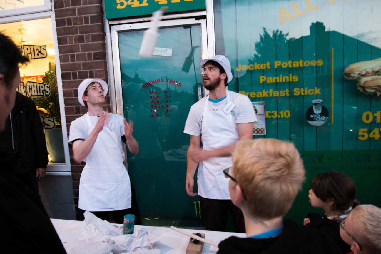 Two performers in white chef's clothes are chucking an object in the air, they are outside a cafe. Three children watch intently from the audience.