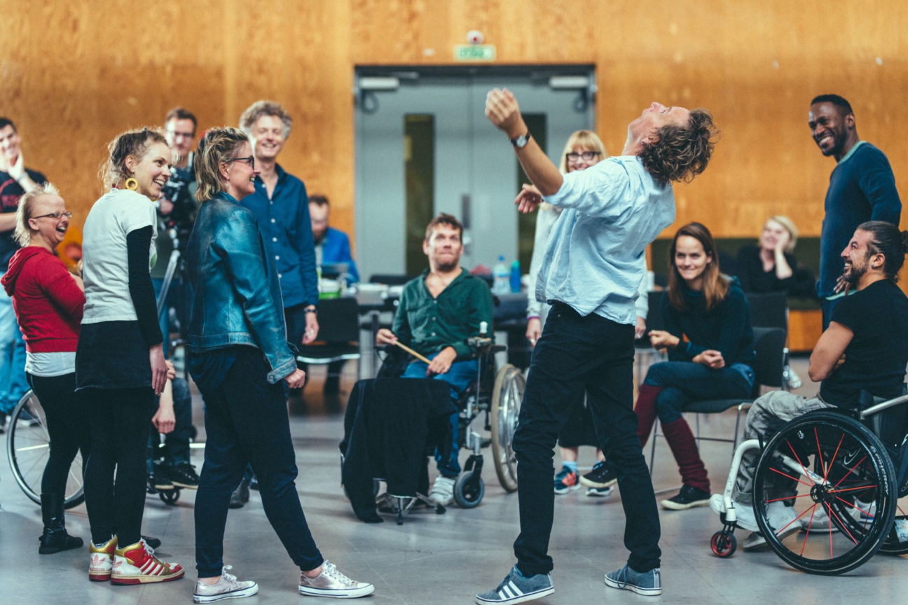 Performer throws his arms and head back animatedly in a circle of performers who are looking at him and smiling.