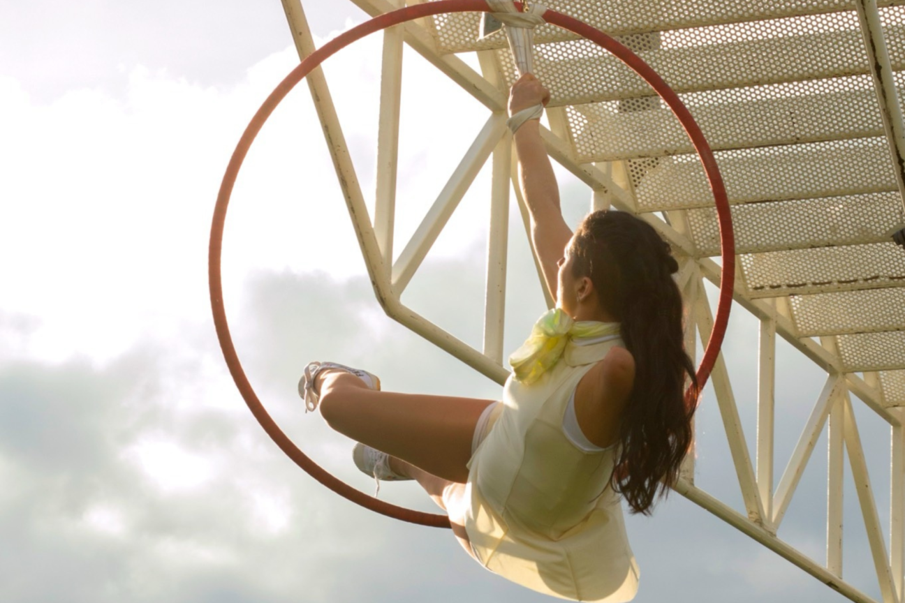 Weighting. Aerialist hangs in red hoop, holding a white strap. She is wearing a yellow, white dress.