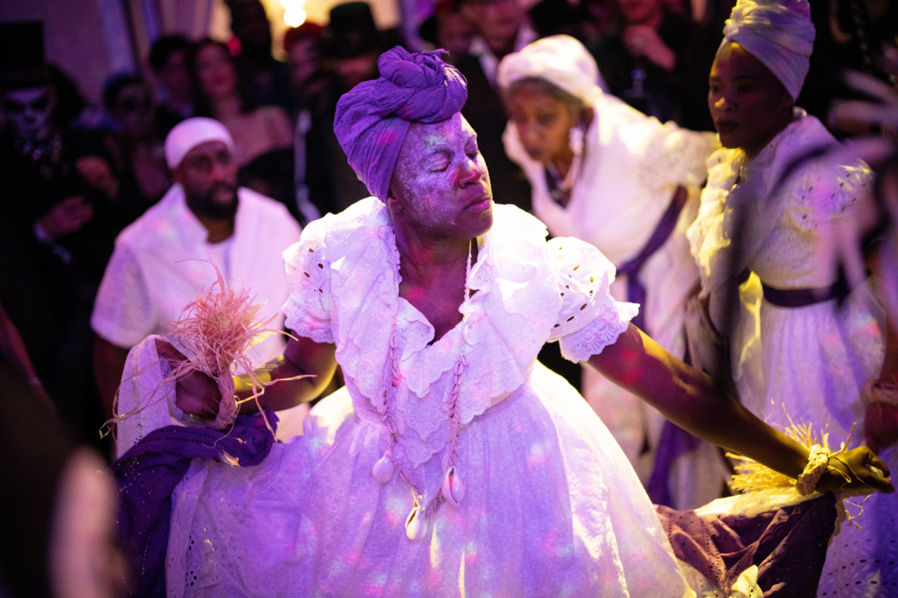 Voodoo Halloween, female performer in purple turban and white frill dress with eyes closed and white face makeup.