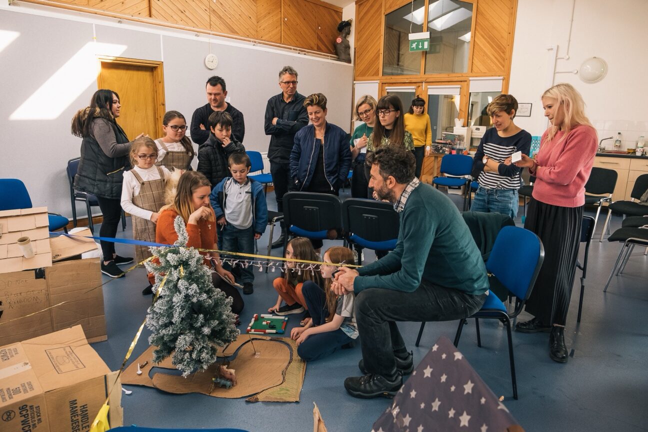 Kitchen Circus Project, 2019. A group of kids stand and sit looking at a small Christmas tree stood on some cardboard and some other small objects on the floor. A man sits on a chair looking at the tree opposite a woman who is talking to the kids who are sat down. A group of adults stand behind the kids, behind chairs looking interested by the tree and smiling.