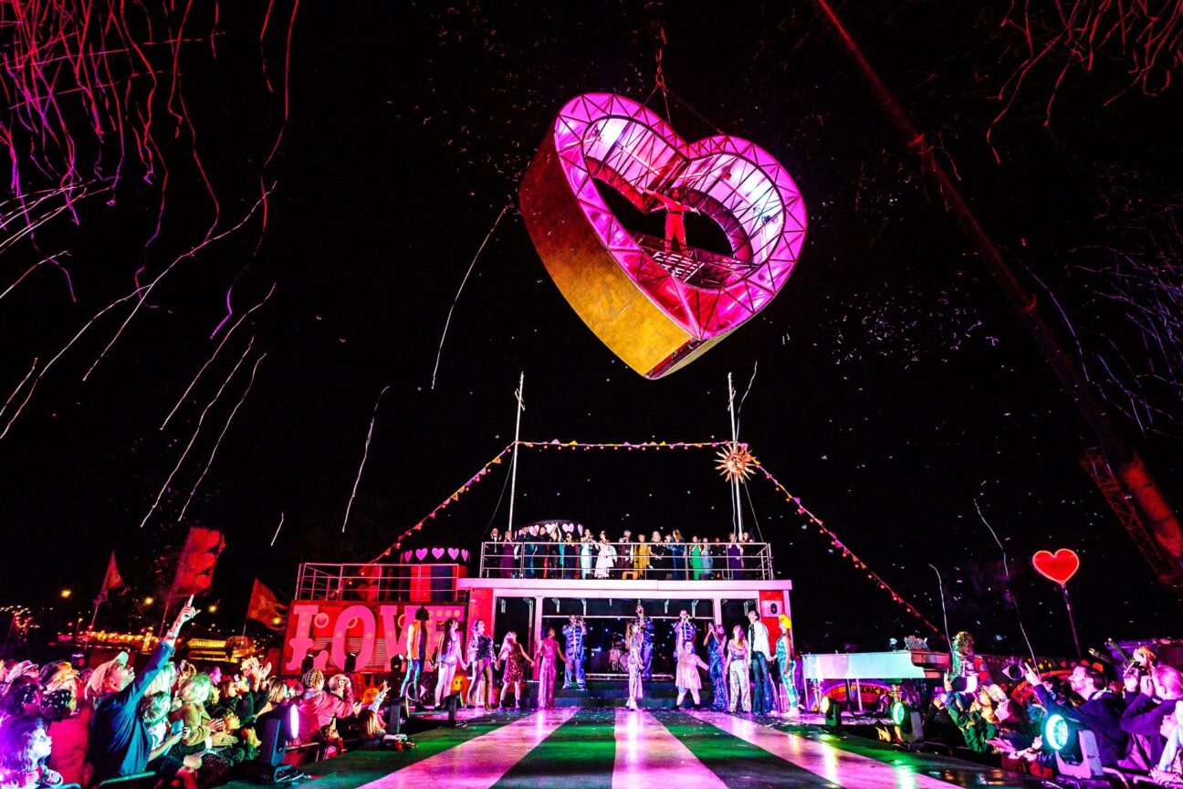 Red heart structure suspended in the air above the Camp Bestival stage. Confetti is released into the air and audience members look up and point.