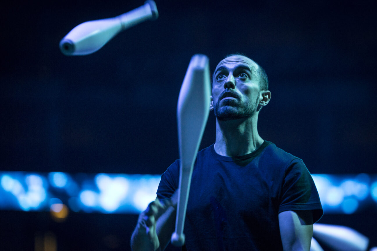 A man concentrates whilst juggling in a darkened room, with a lit screen in the background