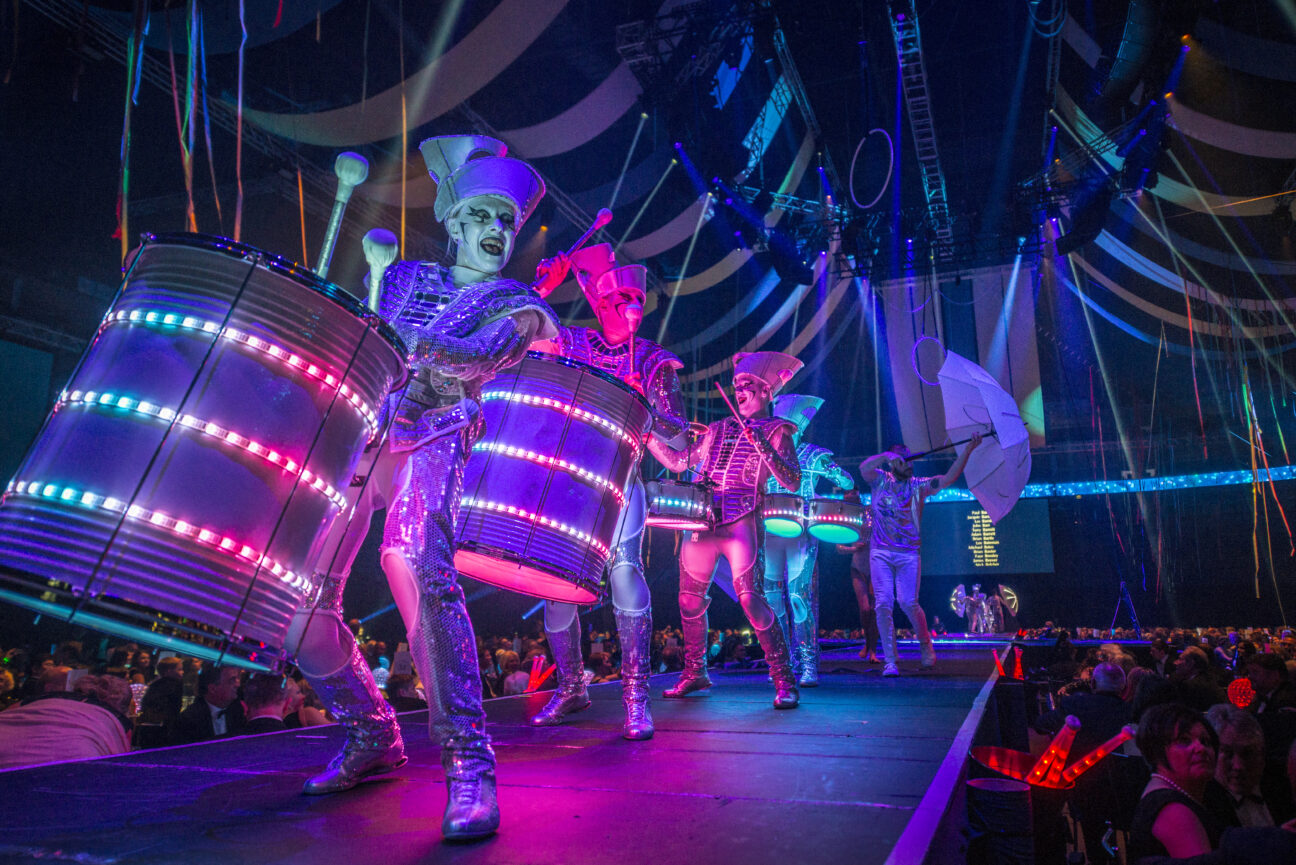 LED Drummers perform down a catwalk, lit in pinks purples and greens and looking at a crowd beyond the frame of the shot