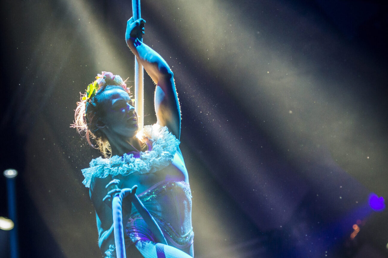 An aerialist clings to her rope, in a darkened room with lights shining down and around her