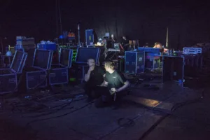 In a darkened room surrounded by technical equipment and wires two tech crew members talk together