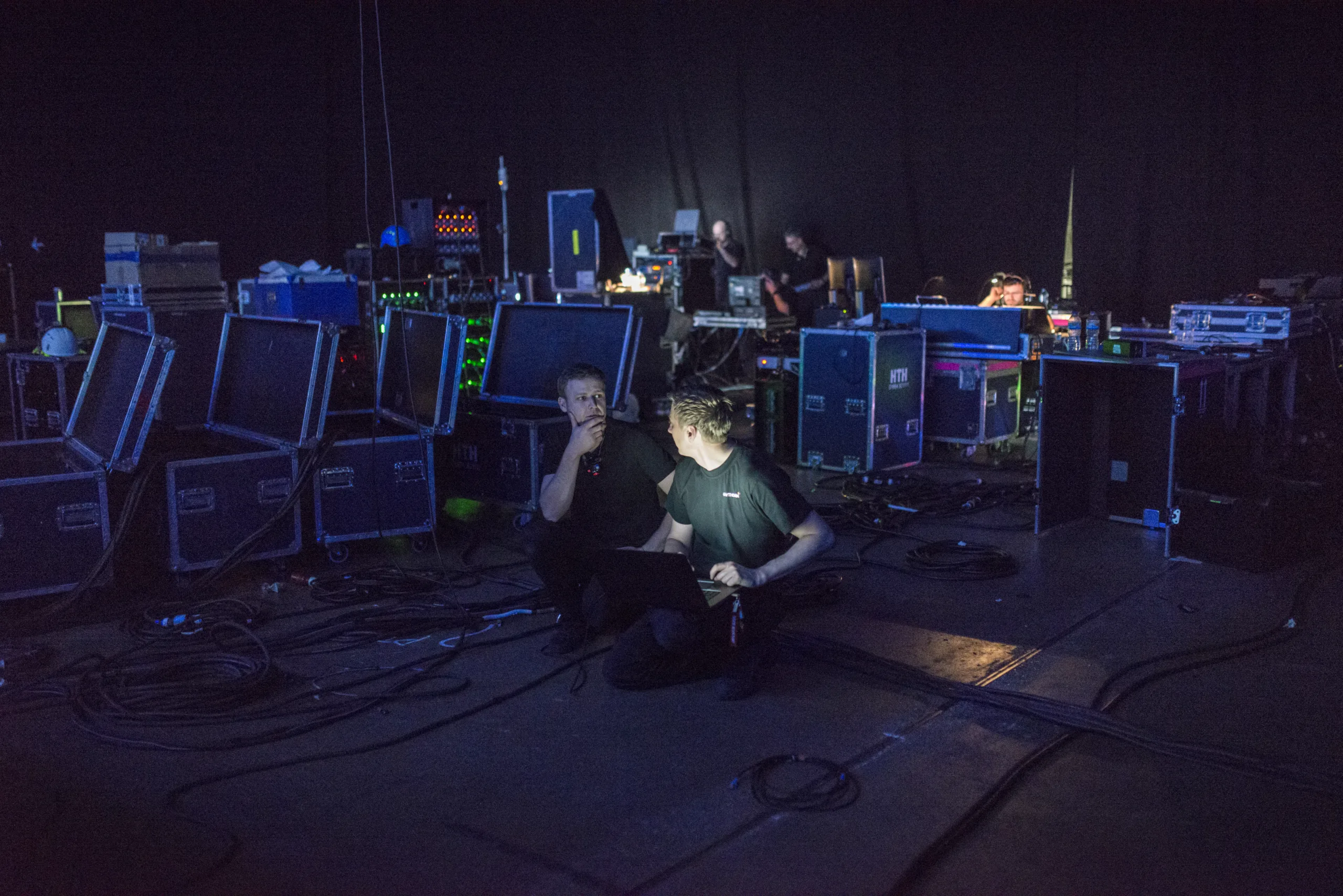 In a darkened room surrounded by technical equipment and wires two tech crew members talk together