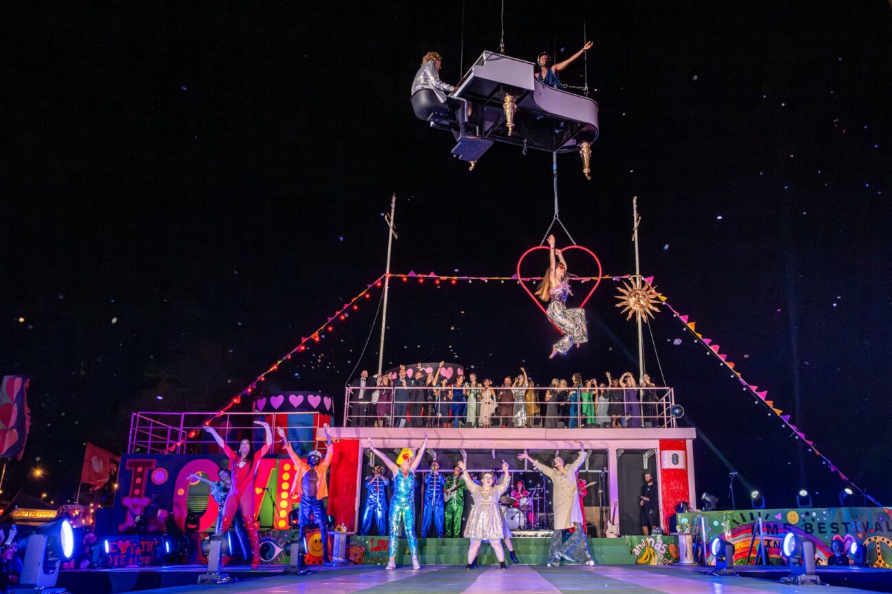 A flying piano and singer raised in the air with an aerialist attached underneath in a heart-shaped aerial hoop. Performers at camp bestival dorset have their arms raised below them on the stage.