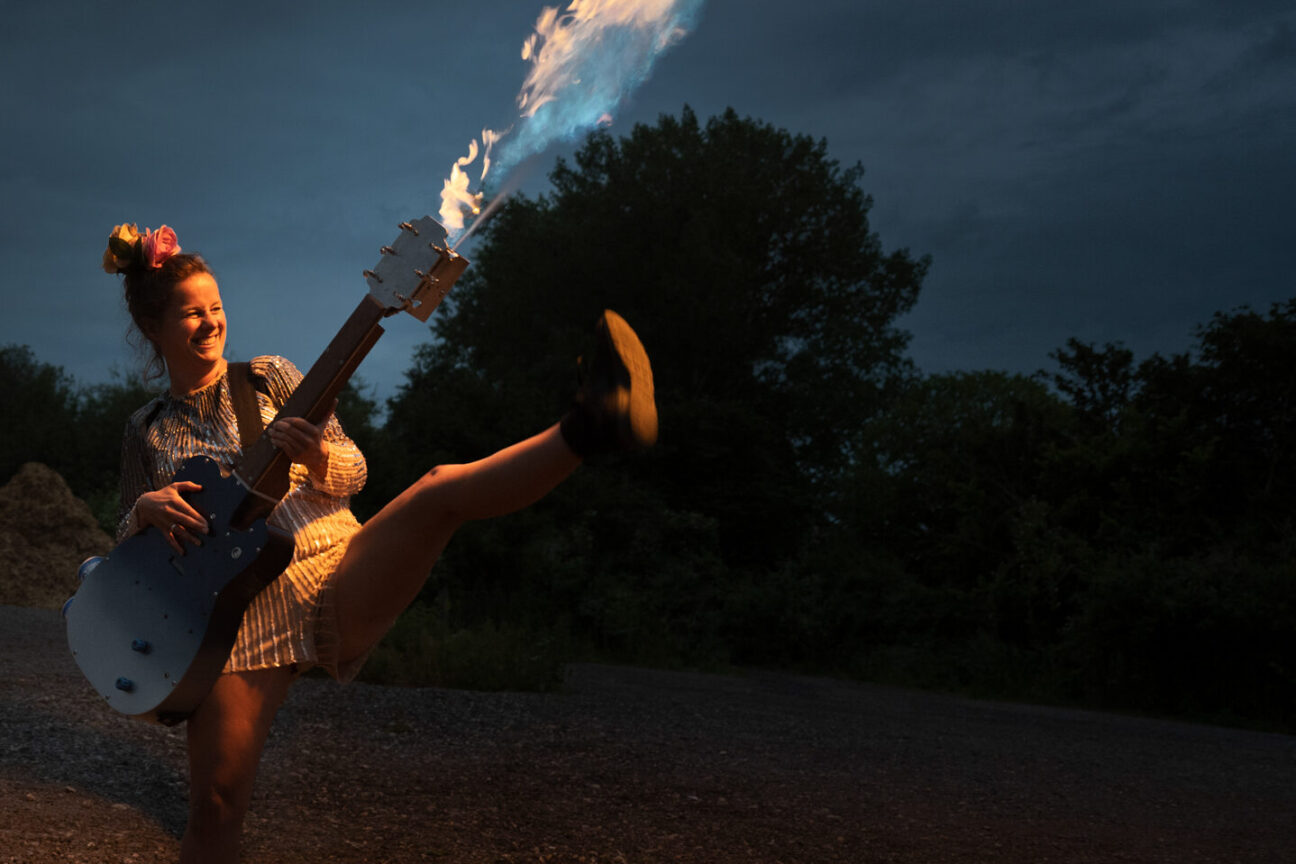 A female guitarist has fire coming out the top of her guitar, she is kicking her leg up in excitement and smiling.